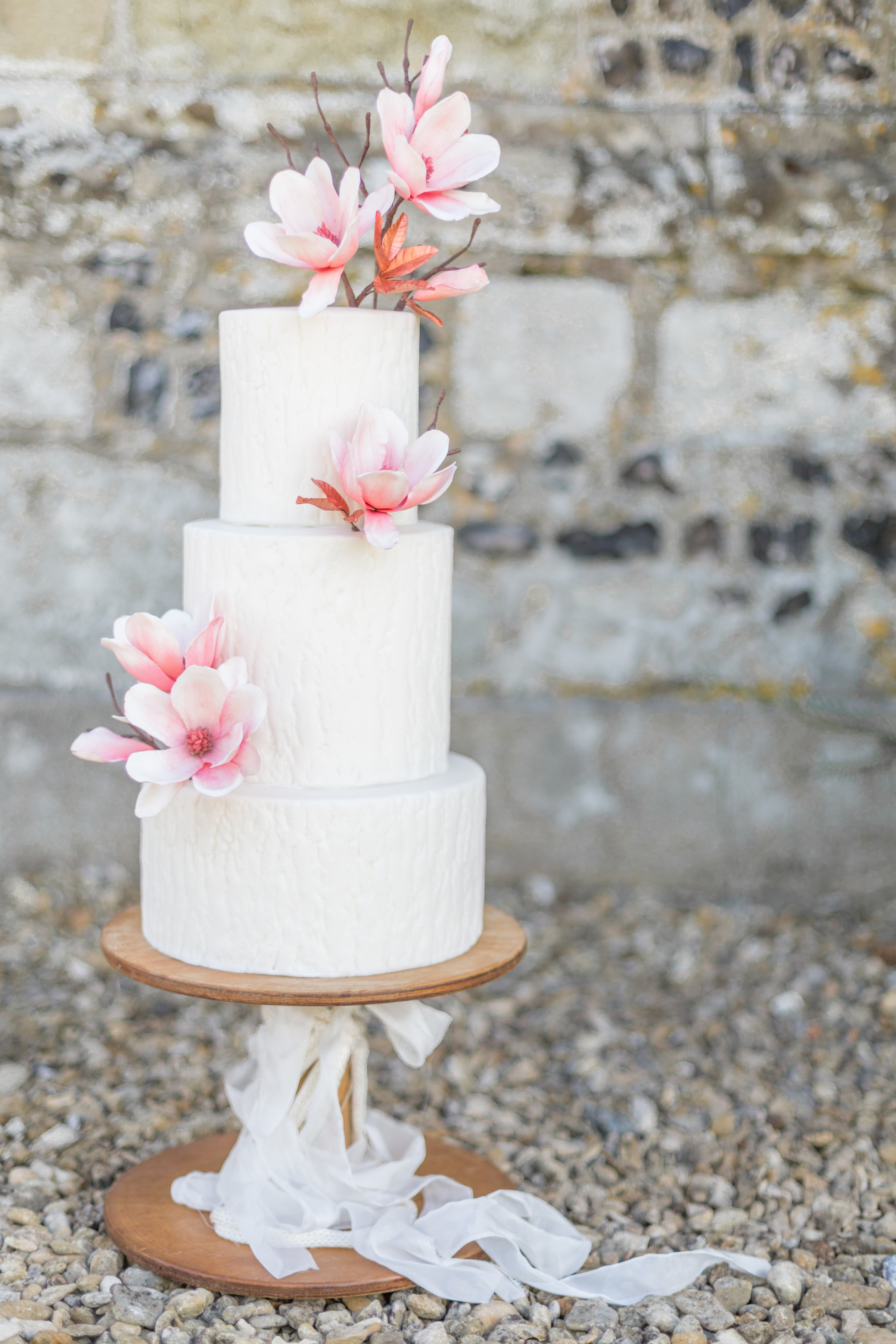 a white textured 3 tier wedding cake with magnolia sugar flowers and rose gold foliage on a wooden cake stand with trailing ribbon