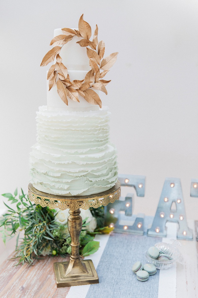 a white and pale green ombre 3 tier wedding cake with delicate pale green ombre ruffles and a gold foliage wreath on a gold cake stand on a table with pale blue table runner and pale blue macarons