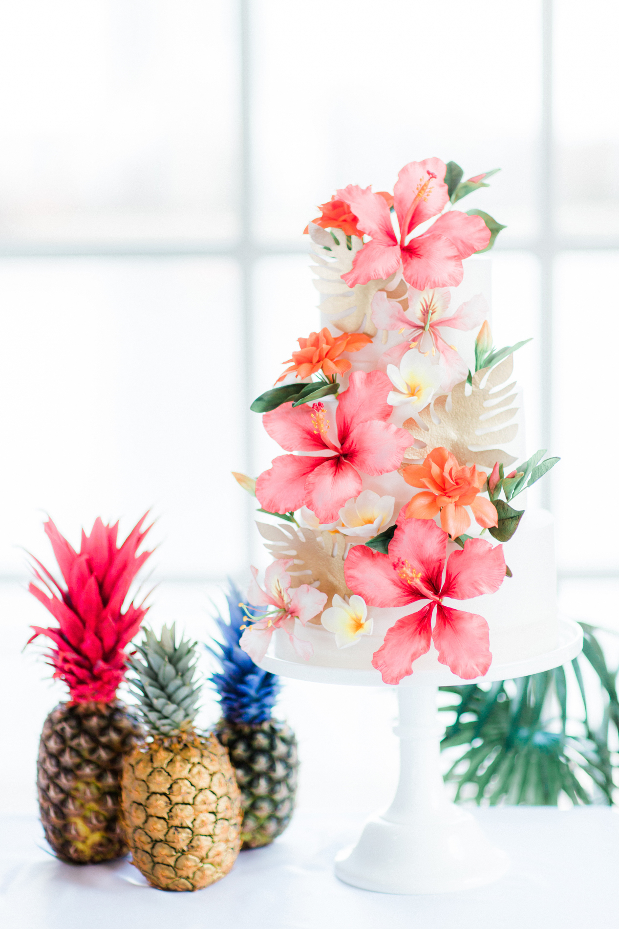 a white 3 tier wedding cake with tropical sugar flowers: pink hibiscus, orange gardenia, white frangipani on a white milk glass cake stand next to coloured pineapples at the west reservoir centre wedding venue hackney