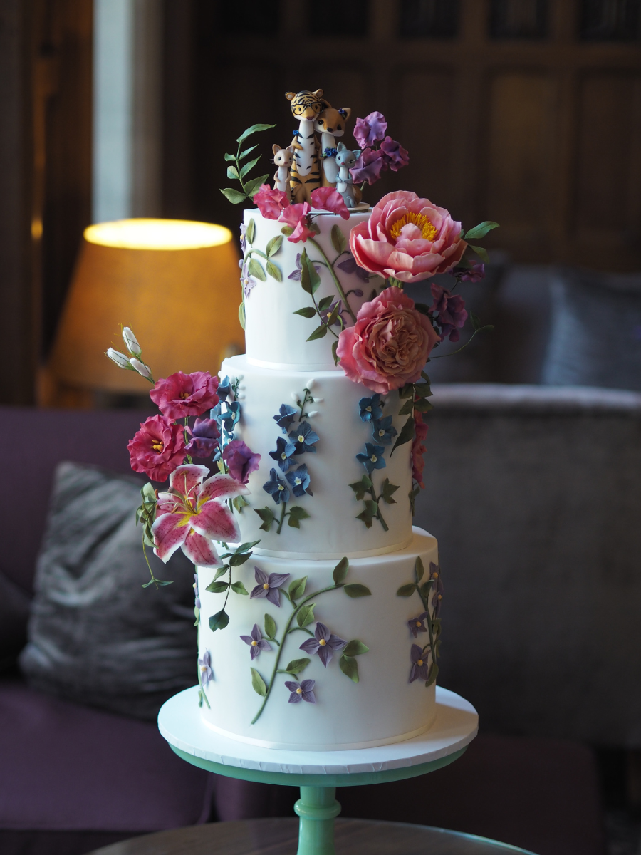 a 3 tier white wedding cake with lots of florals, 2 statement sugar flower arrangements with coral charm peonies, pink english garden rose, pink lisianthus, pink stargazer lily, purple and pink sweet peas and trailing jasmine foliage
