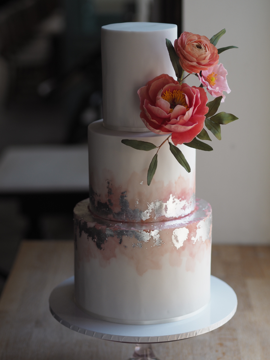 a 3 tier white wedding cake with blush watercolour effect and sliver leaf with a statement sugar flower arrangement of coral charm peony, peach ranunculus and pink japanese anemone and eucalyptus nicholii foliage at the Lordship Pub Dulwich