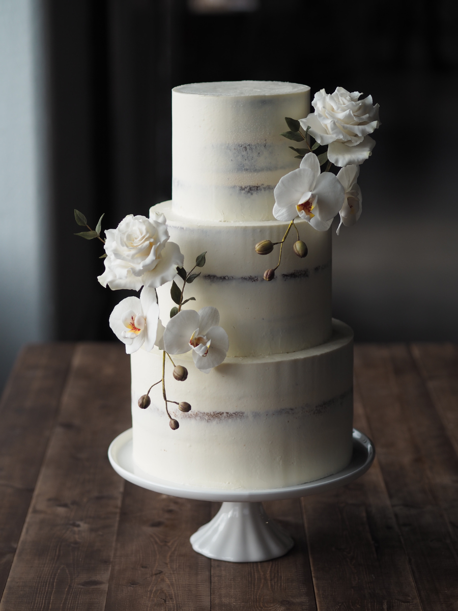 a 3 tier semi-naked wedding cake with 2 statement sugar flower arrangements of white moth phalaenopsis orchids, white roses and eucalyptus parvifolia at trinity buoy wharf wedding venue the chainstore