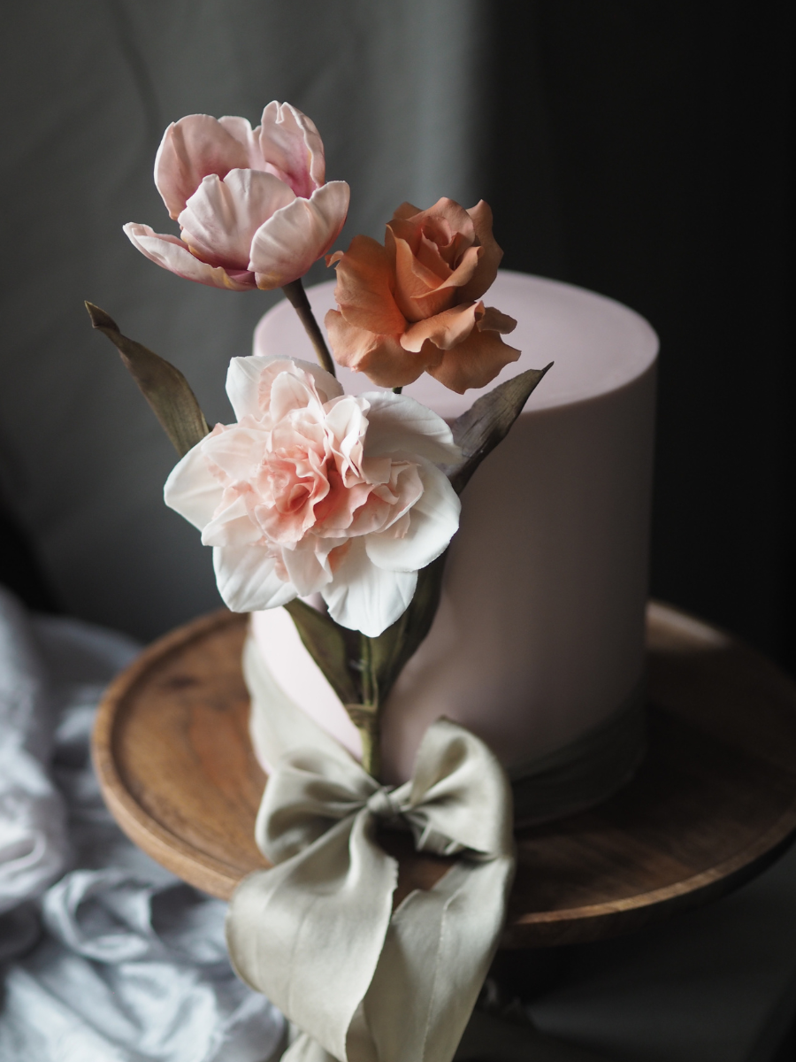 A blush pink single tier cake with grey green silk ribbon and statement sugar flower arrangement of pink tulip, toffee reflexed rose and narcissus replete