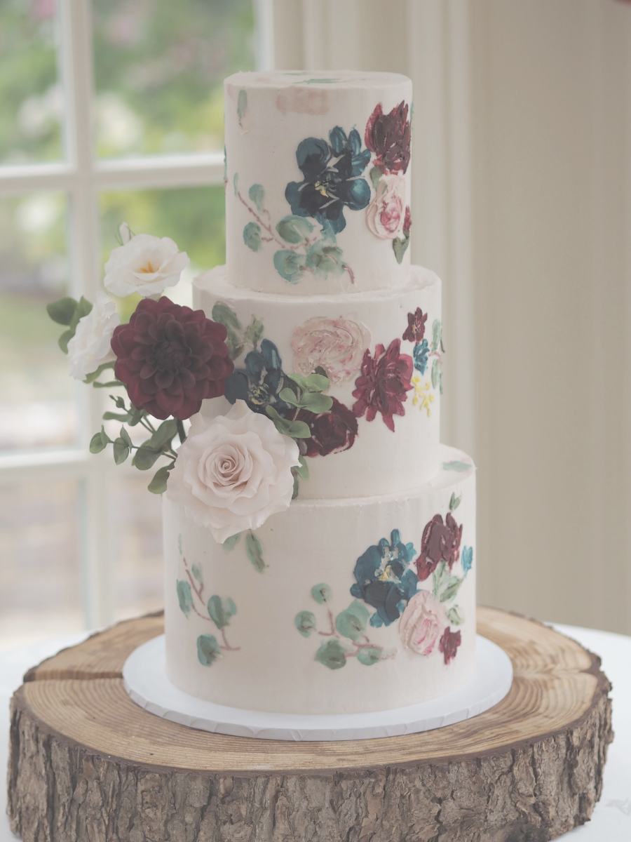 a 3 tier buttercream wedding cake with painted buttercream effect in burgundy, navy blue, blush pink and eucalyptus green with statement sugar flower arrangement of burgundy dahlia, blush rose, white lisianthus and eucalyptus at oaks farm weddings