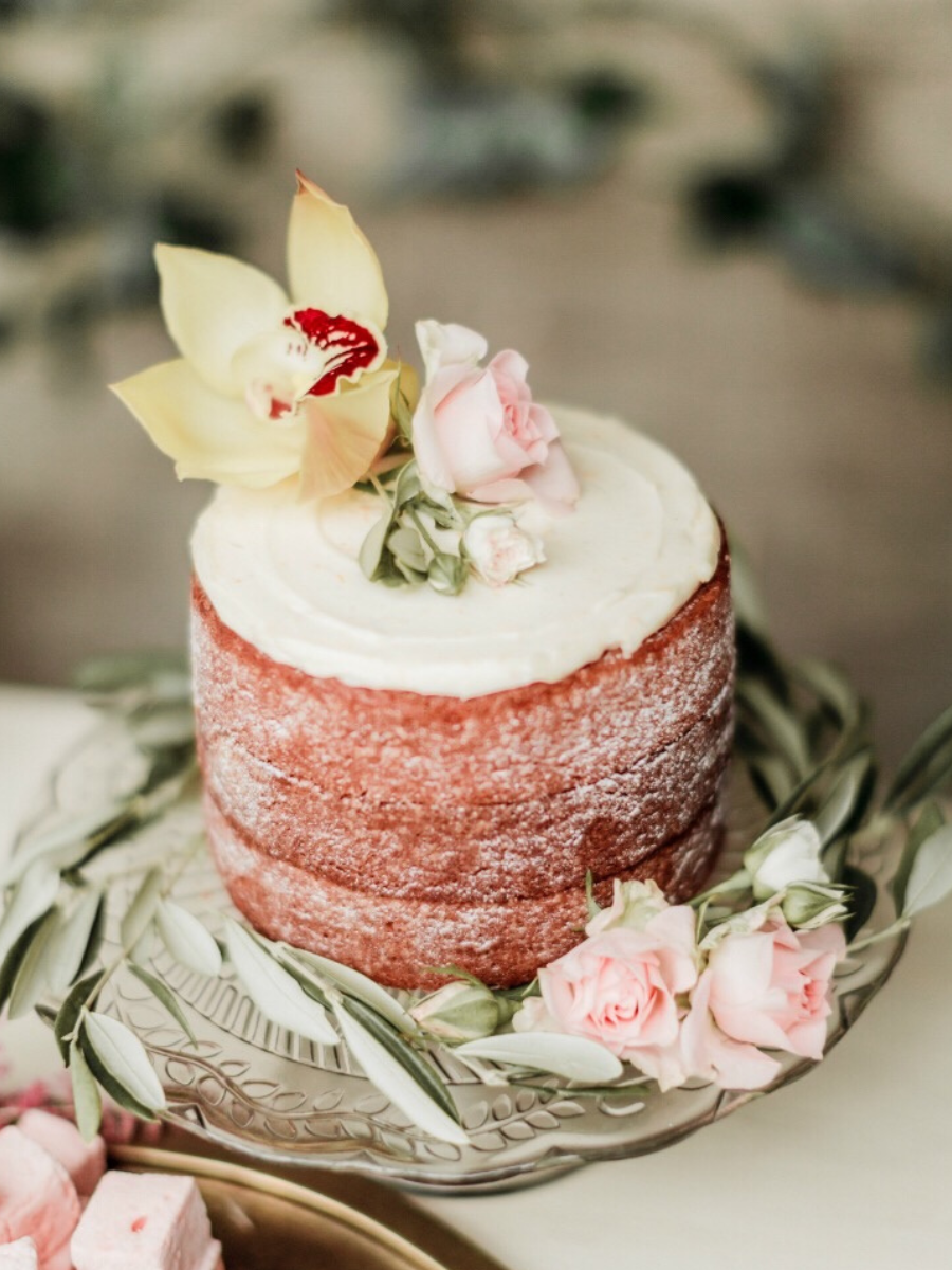 a single tier naked cake dressed with fresh pink and yellow flowers, olive foliage and pink marshmallows